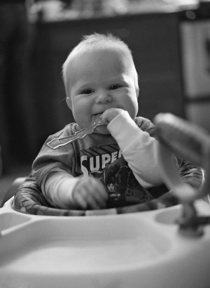 Documentary portrait of a baby boy with a spoon in his mouth sitting in a walker in the kitchen. Shot on a medium format camera using black and white film.