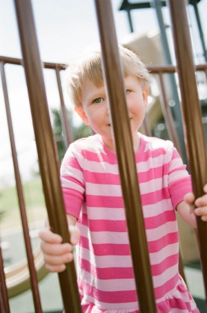 A two year old girl peers through bars on the playground at Nottingham Park in Westminster, CO. Photograph from a documentary portrait session using Fuji 400H film in a Nikon N90s camera.