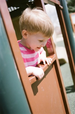 A two year old girl looks through a playhouse window on the playground at Nottingham Park in Westminster, CO. Photograph from a documentary portrait session using Fuji 400H film in a Nikon N90s camera.